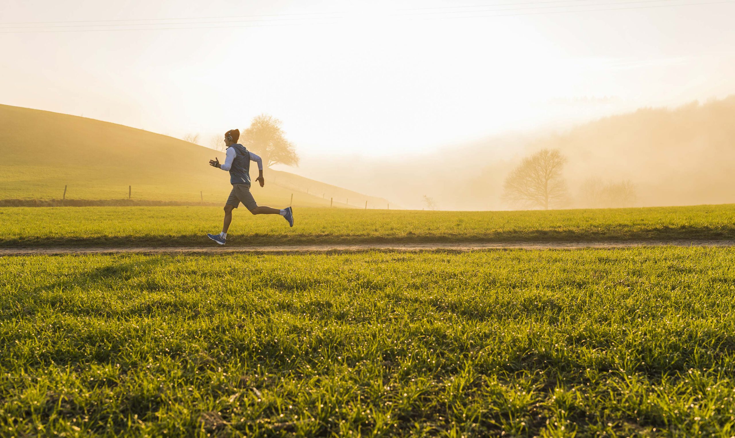 Ein Mann joggt in sportlicher Kleidung durch die Natur. Im Vordergrund ist eine Wiese zu sehen. Im Hintergrund sanfte grüne Hügel mit Gras und Bäumen.