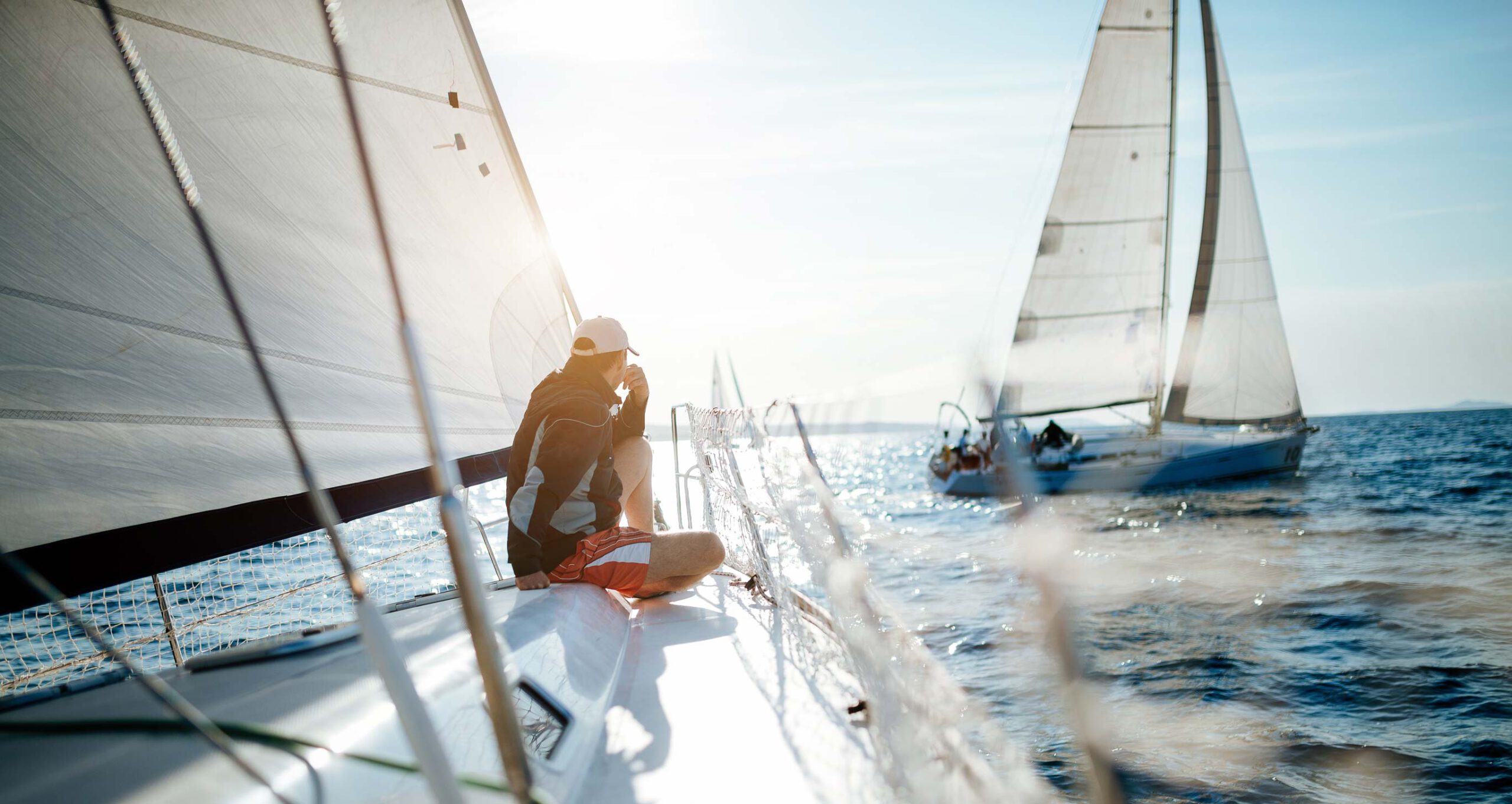 CVM_Segel_setzen Auf einem Gewässer treiben zwei Segelboote. Auf dem einen Boot sitzt ein Mann in entspannter Haltung. Auf dem anderen Boot im Hingerund sind mehrere Menschen.