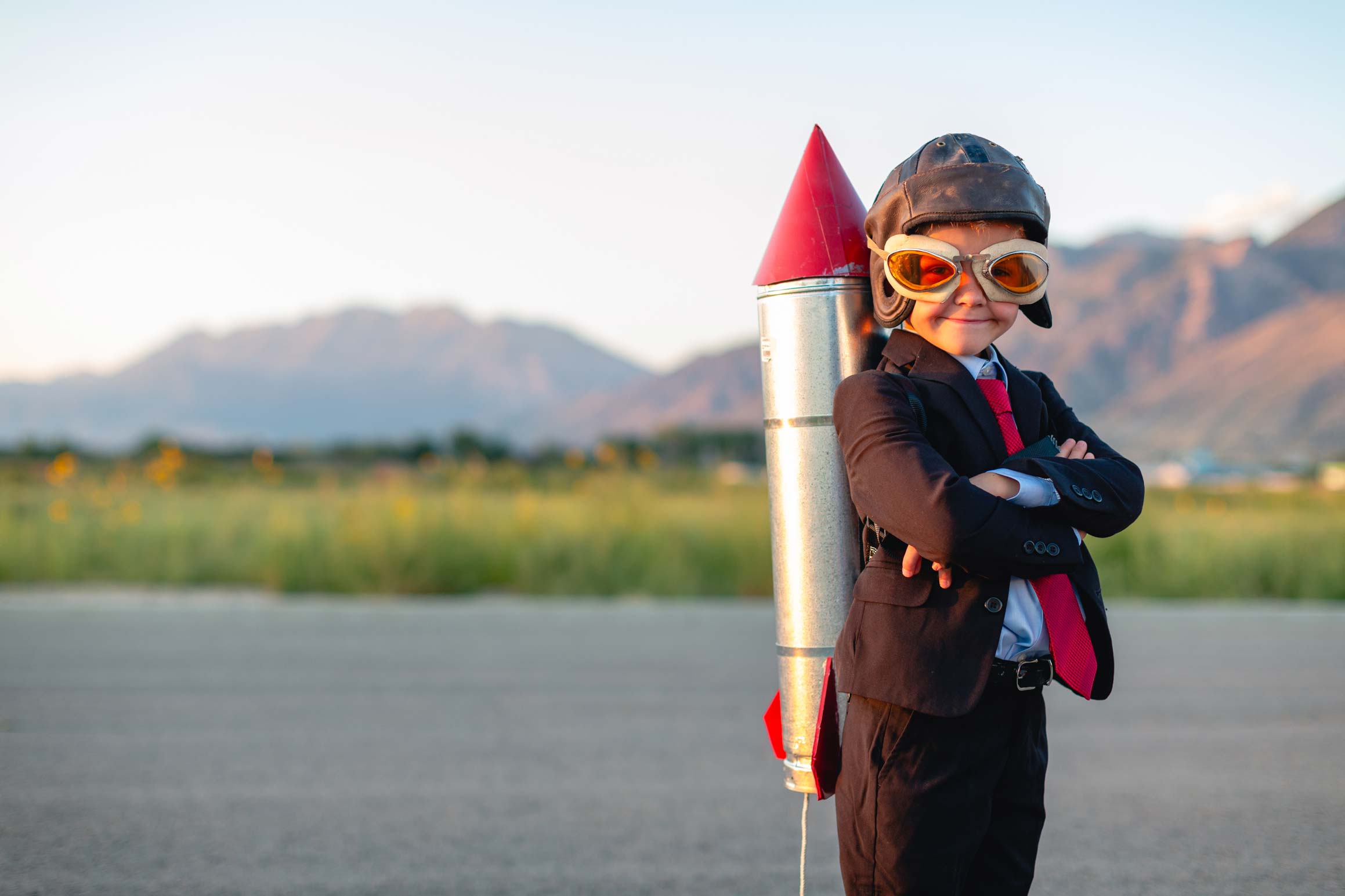 Ein Junge blickt lächelnd in Richtung Kamera. Auf dem Kopf trägt er eine Lederkappe und eine zu große Fliegerbrille. Auf dem Rücken hat er eine Spielzeugrakete.