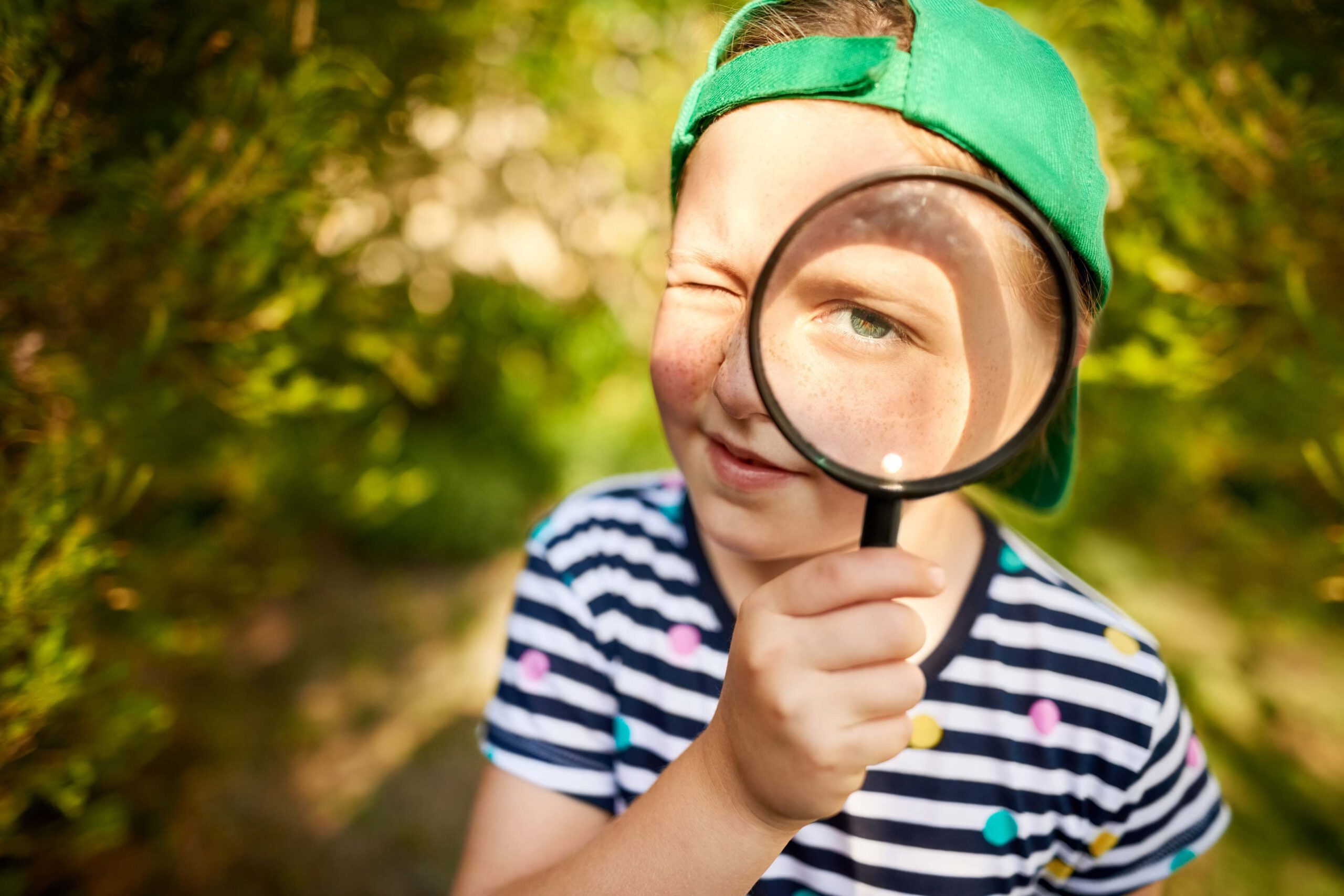 Ein Junge steht dicht vor der Kamera. Er trägt ein Basecape und ein gestreiftes T-Shirt und hält sich eine Lupe dicht vor das linke Auge.